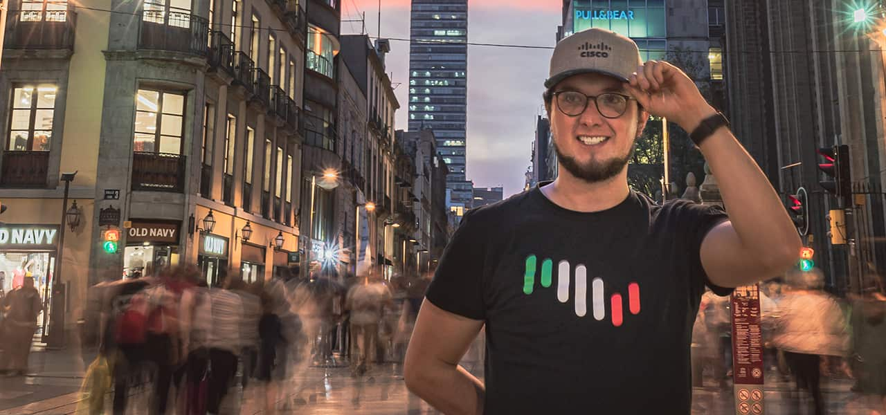 Male wearing a Cisco Hat and shirt holding left hand to his hat on a street in Mexico City